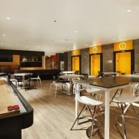 room with yellow walls, shuffleboard on side and tables with white chairs on modern bar wood flooring by Dralynn Floor Covering Inc.
